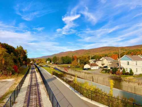 A scenic view of a railway track beside a canal, with colorful autumn trees and houses against a backdrop of mountains.