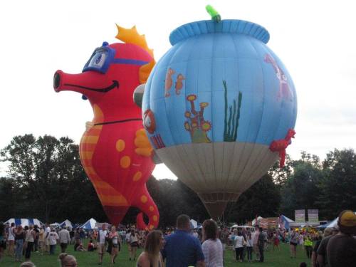 Colorful hot air balloons shaped like a seahorse and a fishbowl, surrounded by a crowd at an outdoor event.
