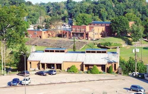 Aerial view of a brick building with a metal roof, surrounded by trees and parked cars, near a grassy area and other structures.
