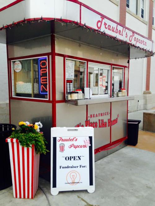 A vintage popcorn stand with a red and white striped design, featuring an "Open" sign and a fundraiser poster outside.