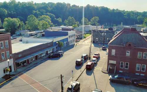 Aerial view of a small town street with parked cars, shops, and trees in the background under a sunny sky.