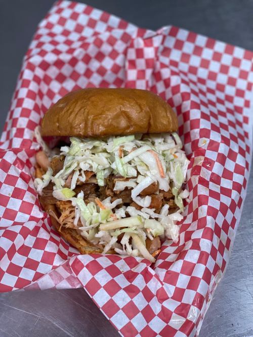 A pulled pork sandwich topped with coleslaw, served in a red and white checkered paper.