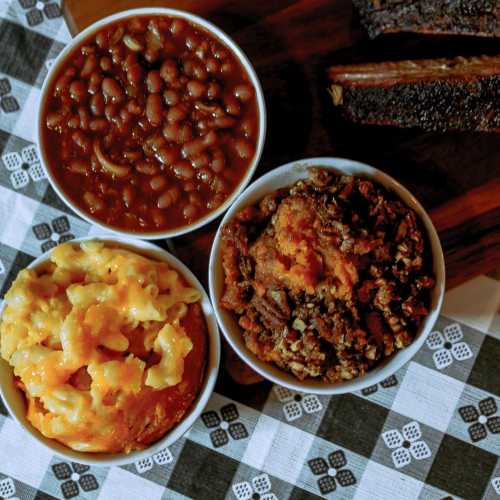 Three bowls of food: baked beans, macaroni and cheese, and a meat dish, on a checkered tablecloth.