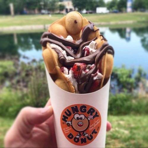 A hand holding a cone filled with bubble waffles, whipped cream, strawberries, and chocolate drizzle by a river.