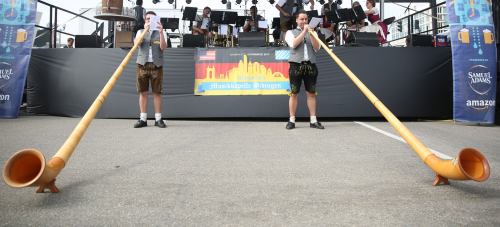 Two musicians in traditional attire play long wooden alphorns on a stage with a band in the background.