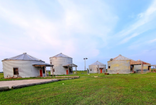 Four unique, round buildings with metal roofs sit on a grassy field under a blue sky.