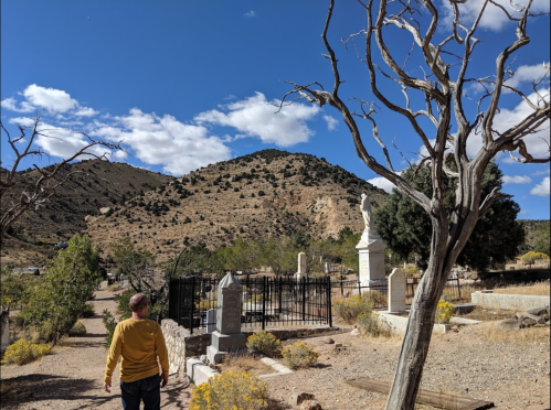 A person walks through a cemetery with dry trees and mountains in the background under a blue sky with clouds.