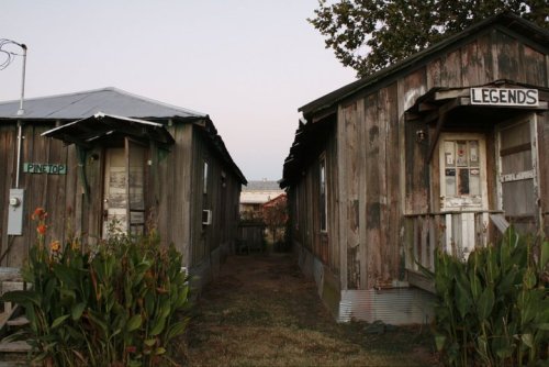Two weathered wooden cabins labeled "PINTOP" and "LEGENDS," separated by a grassy path and surrounded by plants.
