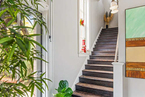 Bright hallway with wooden stairs, large windows, and greenery, leading to an upper level with modern artwork on the wall.