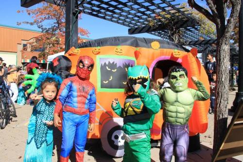 Children in colorful costumes, including superheroes and a dinosaur, pose in front of a decorated Halloween-themed van.