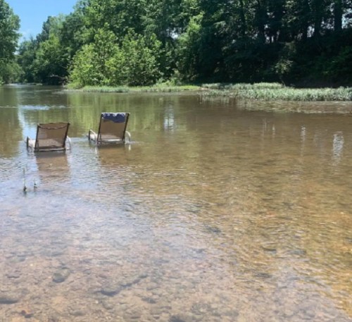 Two empty lounge chairs sit in shallow water by a serene river, surrounded by lush greenery.