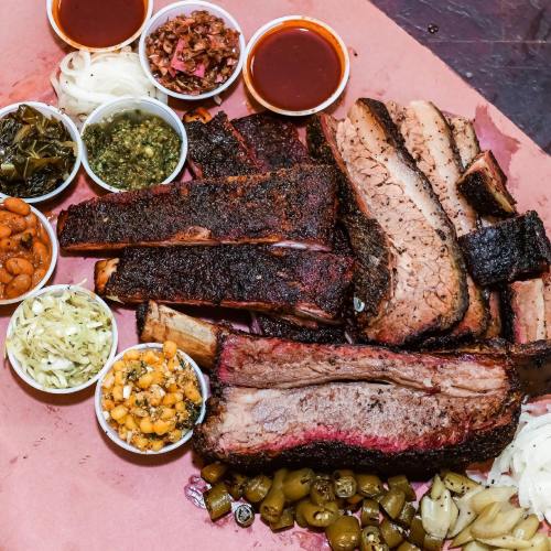 A platter of smoked meats including ribs and brisket, accompanied by various sides and sauces in small bowls.