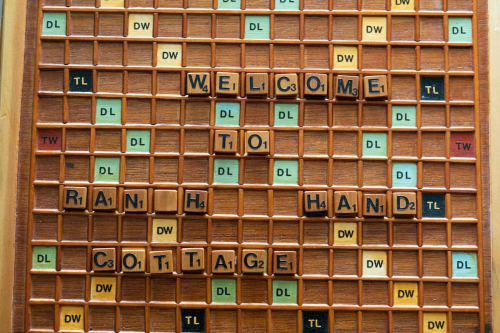A wooden Scrabble board displaying the words "Welcome to Rain Hand Cottage" with various scoring tiles.