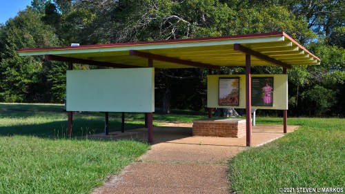 A small, open pavilion with a green roof and brick base, surrounded by grass and trees.