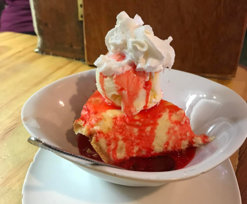 A slice of cheesecake topped with strawberry sauce and a scoop of ice cream, garnished with whipped cream in a bowl.