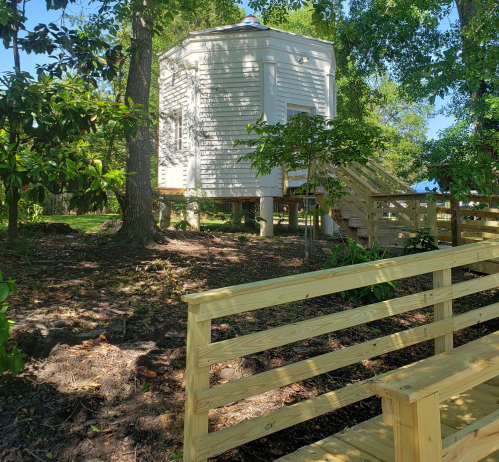A small white building on stilts surrounded by trees, with a wooden bench in the foreground and stairs leading up.