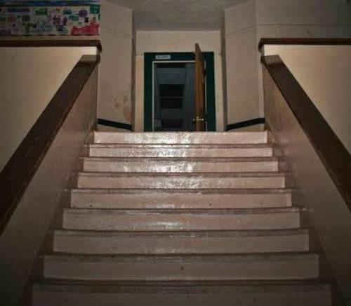 A dimly lit staircase leads to an open door at the top, with walls showing signs of wear and age.