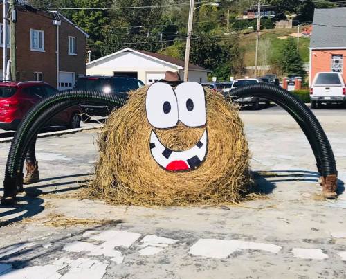 A large hay bale with a smiling face, surrounded by black tubes, set in a parking area with buildings in the background.