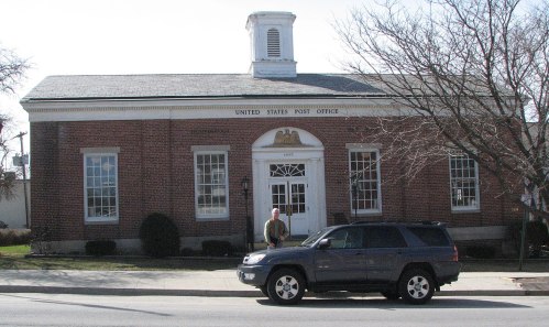 A brick building with a sign reading "United States Post Office," featuring a man standing near a parked SUV.
