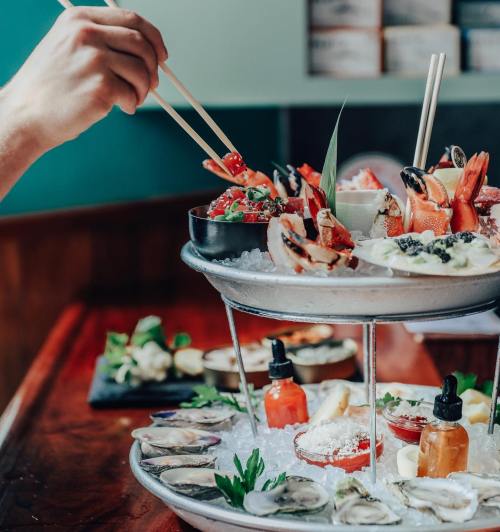 A hand using chopsticks to pick seafood from a tiered platter filled with oysters, shrimp, and garnishes.