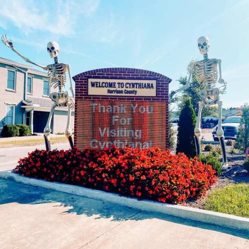Welcome sign to Cynthiana, featuring two skeletons and vibrant flowers in front.