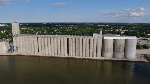 Aerial view of a large grain silo complex beside a calm river, with green fields and a town in the background.