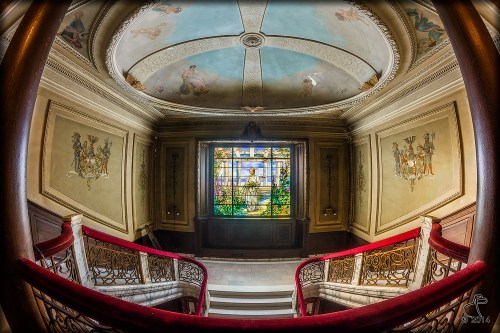 A grand staircase with ornate railings leads to a stained glass window, surrounded by intricate ceiling and wall paintings.