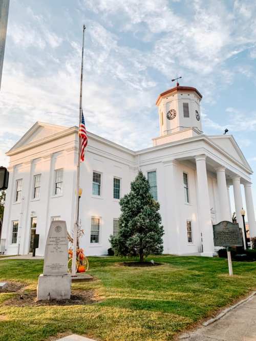 A white historic building with a clock tower, surrounded by greenery and an American flag at half-mast.