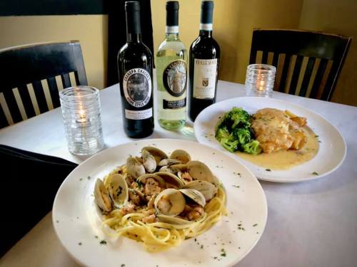 A table set with two plates of pasta and chicken, accompanied by three bottles of wine and candles.