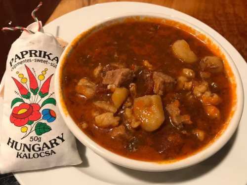 A bowl of hearty stew with meat and vegetables, accompanied by a small bag of Hungarian paprika.