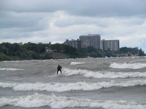 A person surfing on choppy waves with a city skyline in the background under a cloudy sky.