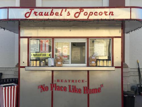A popcorn stand with a red and silver exterior, featuring the name "Traubel's Popcorn" and a slogan "No Place Like Home."