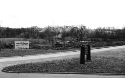 Black and white image of a rural landscape with a sign for a new boat harbor and cleared land in the background.