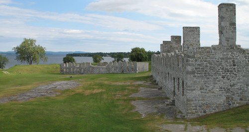Ruins of a stone structure on grassy land by a lake, with trees and mountains in the background under a cloudy sky.