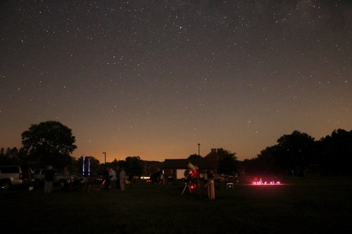A night sky filled with stars above a gathering of people with telescopes in a grassy field.