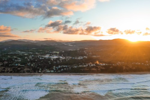 Aerial view of a coastal town at sunset, with waves crashing on the shore and mountains in the background.
