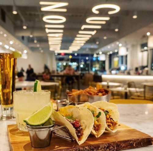 A wooden platter with three tacos, a margarita, and a beer, set in a modern restaurant with bright lighting.