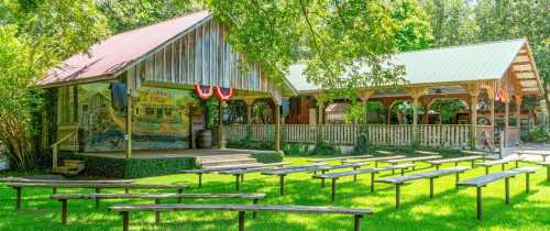 A rustic outdoor venue with wooden structures and benches on a grassy area, surrounded by trees.