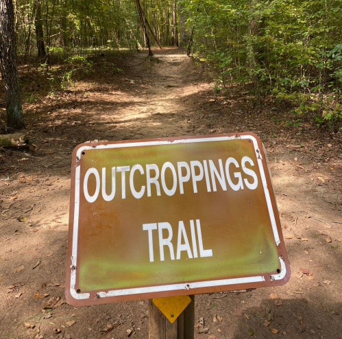 A weathered sign reading "Outcroppings Trail" stands at the entrance of a wooded path.