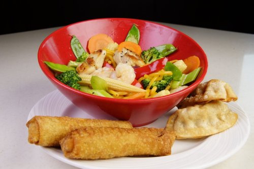 A vibrant bowl of stir-fried vegetables and shrimp, accompanied by crispy spring rolls and dumplings on a plate.