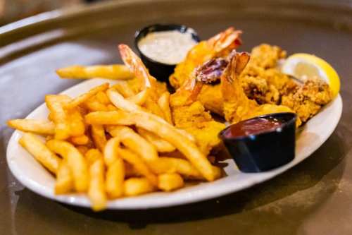 A plate of golden fried shrimp and chicken with French fries, served with dipping sauces and a lemon wedge.