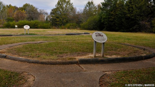 A grassy area with circular pathways and informational signs among trees under a cloudy sky.