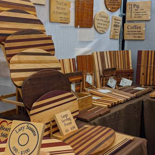 A display of wooden cutting boards and serving pieces with various designs and price tags at a craft fair.