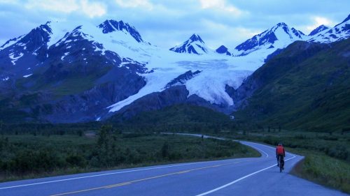A cyclist rides along a winding road with snow-capped mountains and a glacier in the background during twilight.