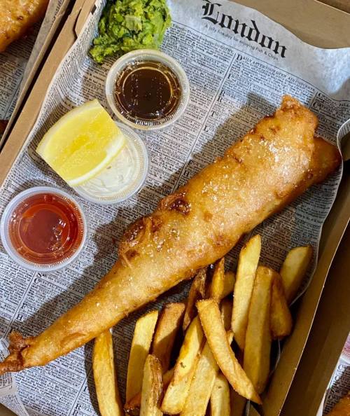 A large piece of fried fish with fries, lemon, and dipping sauces on a newspaper-lined tray.