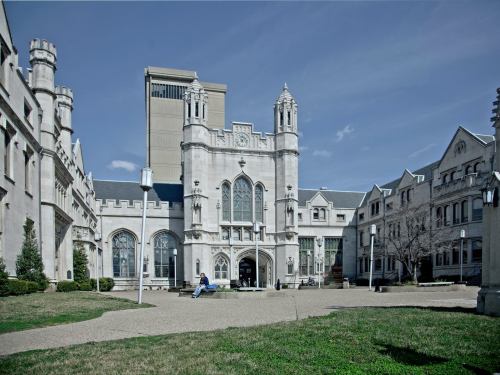A historic stone building with gothic architecture, surrounded by grass and trees, under a clear blue sky.