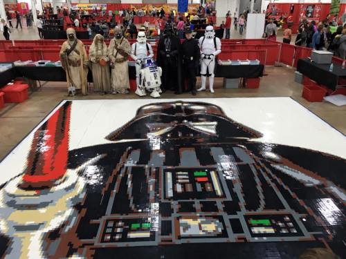 A large LEGO mural of Darth Vader with costumed characters, including Stormtroopers and R2-D2, at an event.