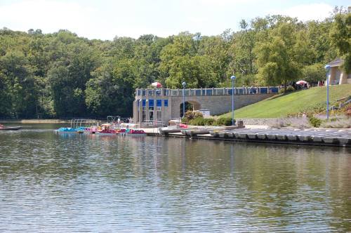 A serene lake scene with a boathouse, paddle boats, and lush greenery in the background under a clear blue sky.