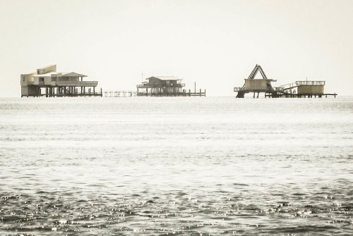 Three structures on stilts stand in calm waters, shrouded in a soft, hazy light.