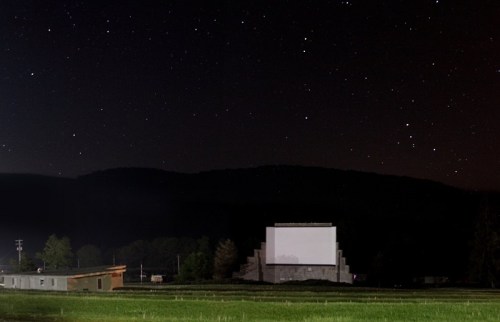 A dark sky filled with stars above a drive-in movie screen and grassy field, surrounded by distant hills.
